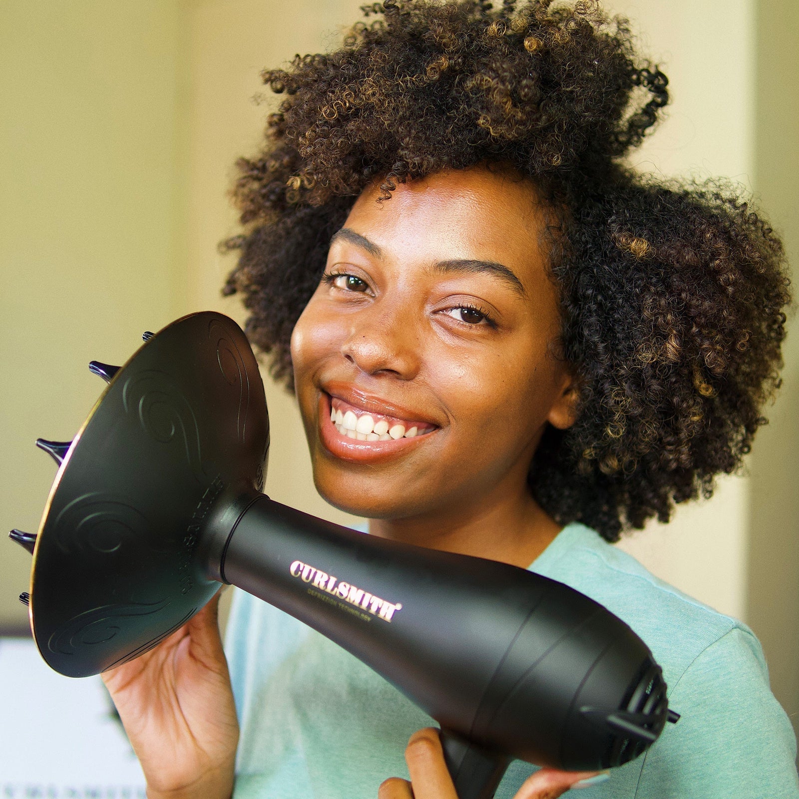 Person holding a black hair dryer with 'Curlsmith' branding, smiling.
