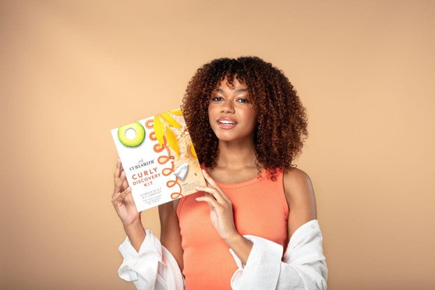 woman with curly hair holds up the Curlsmith discovery kit box against a faded orange backdrop